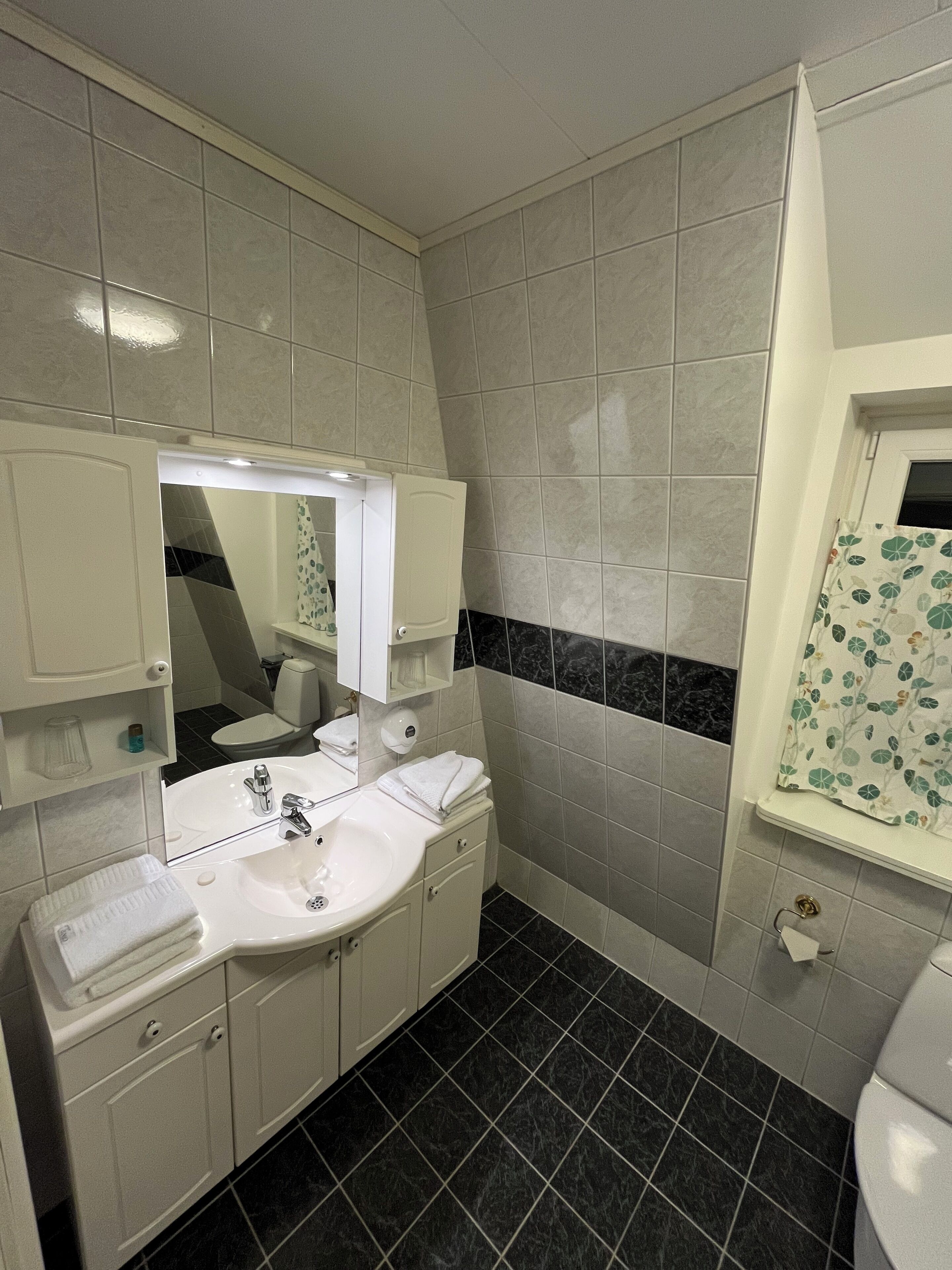 Une salle de bain avec un lavabo blanc, un miroir et des toilettes. Le sol est recouvert de carreaux noirs et blancs. Il y a une fenêtre avec un rideau.