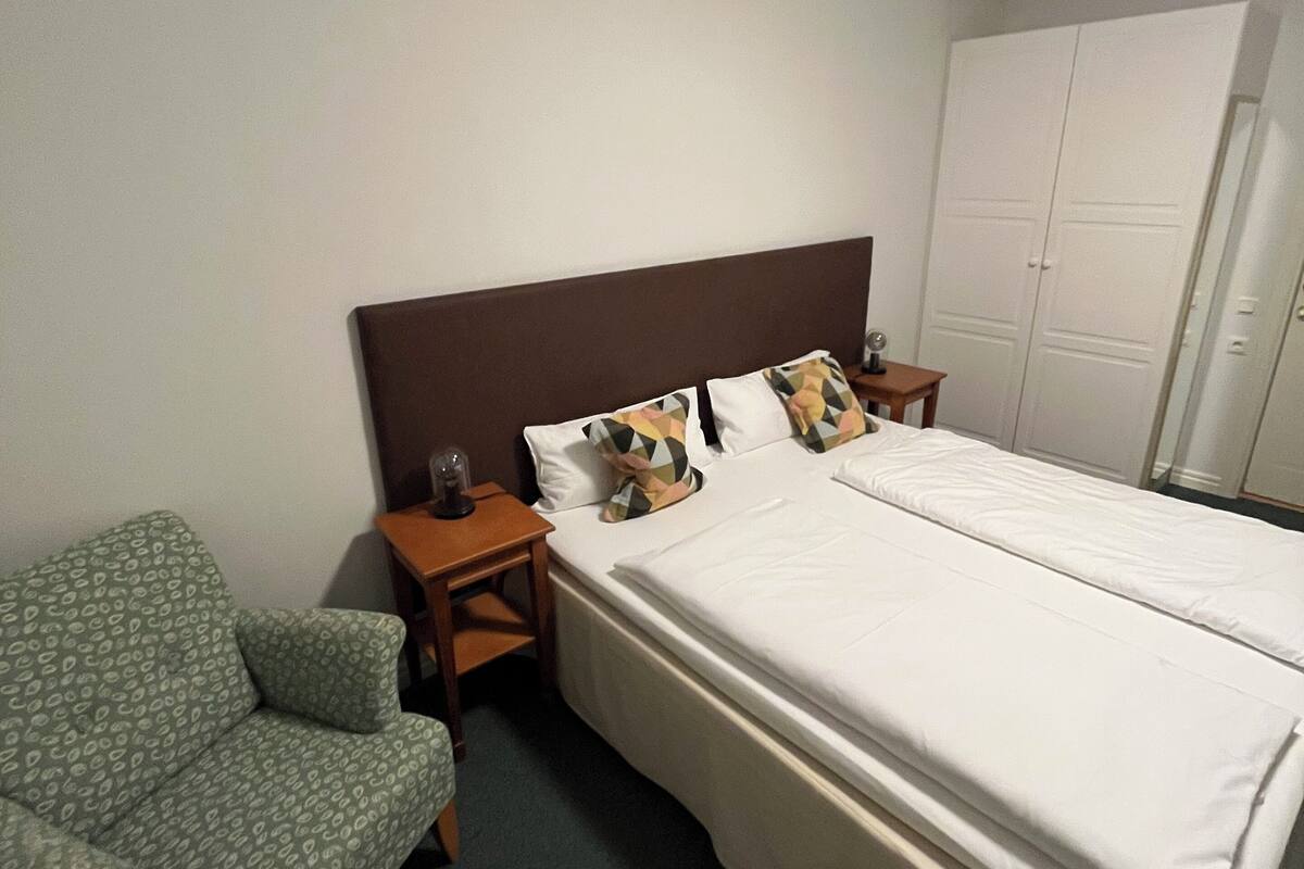 Quarto de hotel com uma cama, uma cadeira, uma mesa de cabeceira e um guarda-roupa.