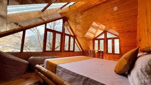 A cozy wooden cabin bedroom with a large bed, a sitting area with a chair, and a view of the outdoors through large windows. - Virman Riverside Resort (Banjar)