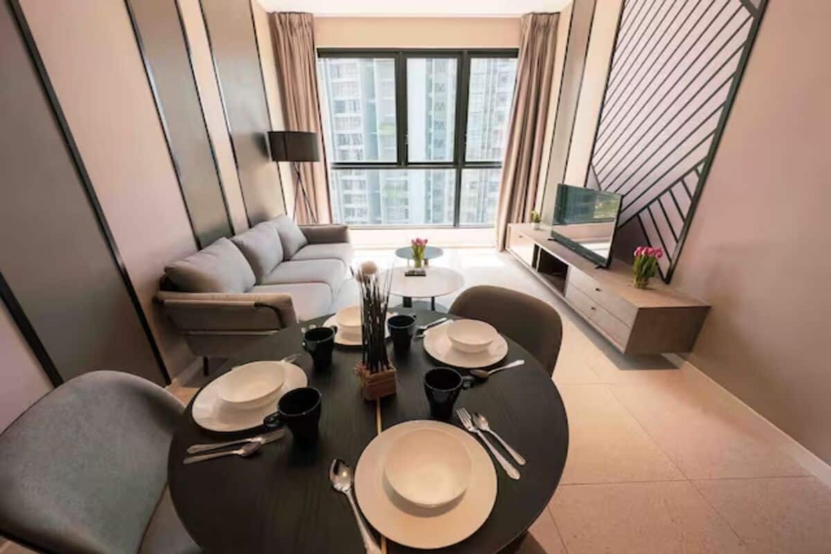 Una sala de estar moderna con una mesa de comedor dispuesta para cuatro personas, un sofá, un televisor y un ventanal con vistas a la ciudad.