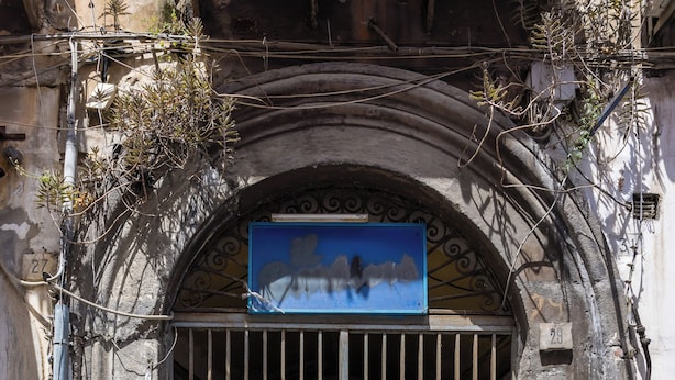 A weathered metal gate with a blue sign above it, set within an arched stone doorway. - Nefelu apartments (Naples)