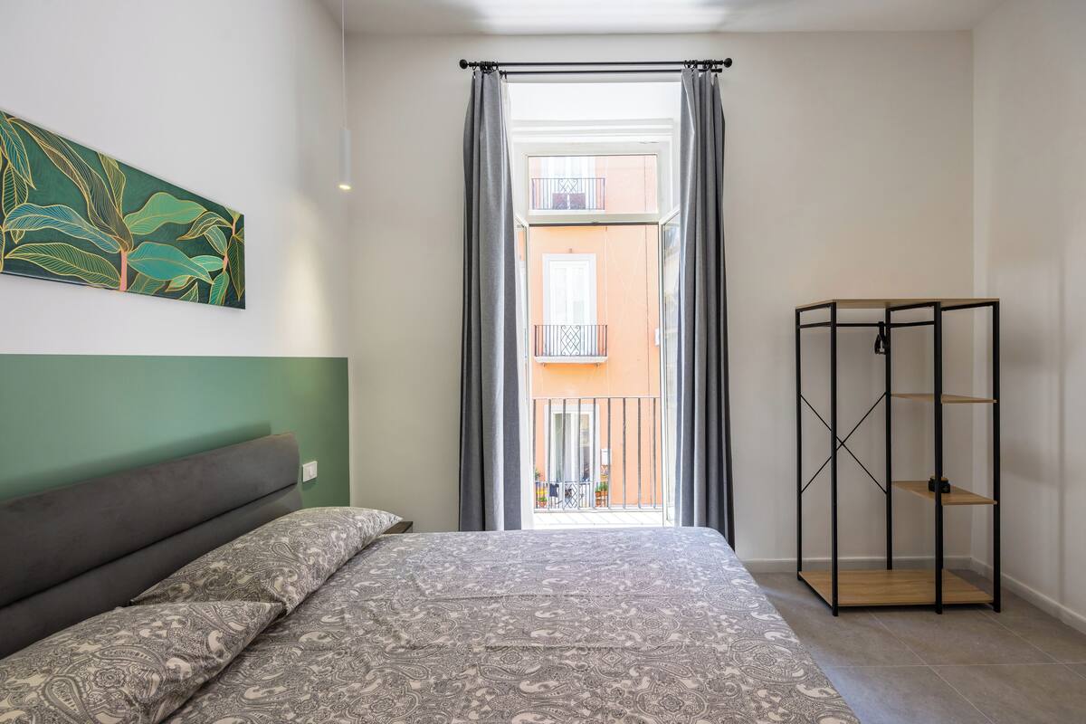 A bedroom with a bed, a green wall, a window with curtains, and a balcony.