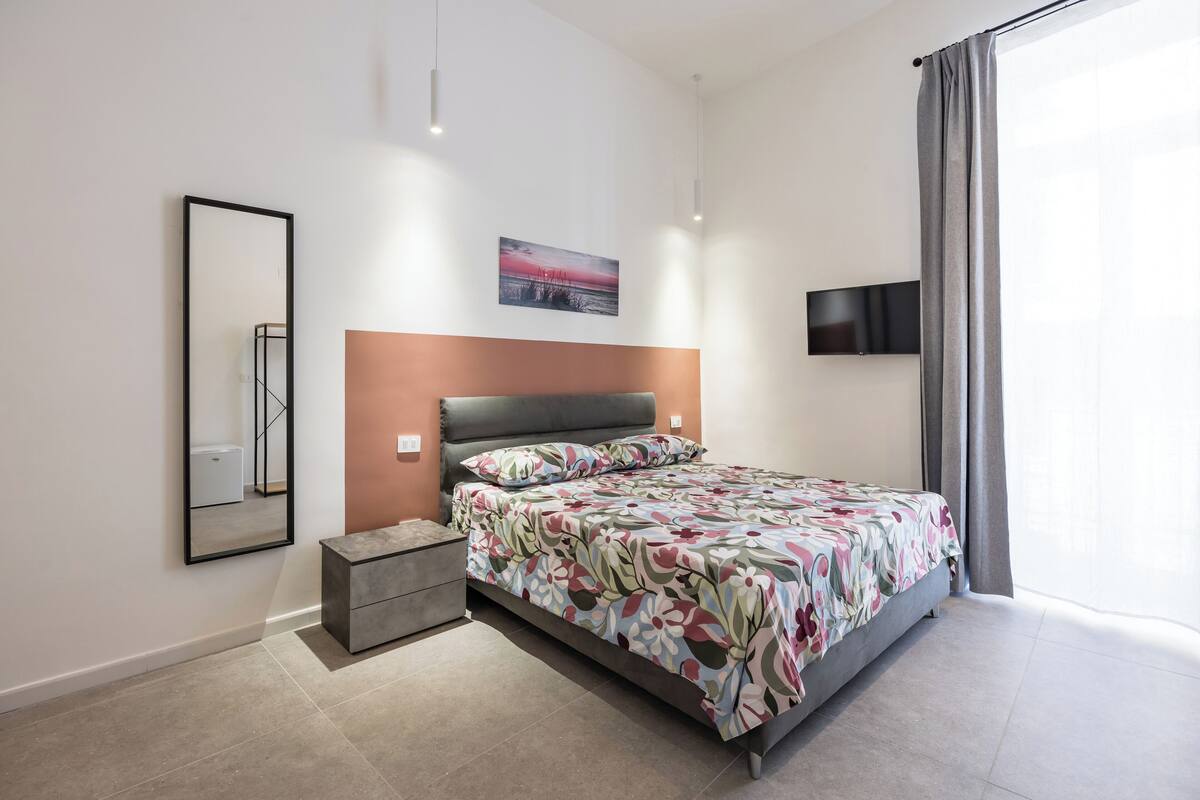 A modern bedroom with a large bed, a nightstand, a small dresser, a wall-mounted TV, and a painting above the bed.