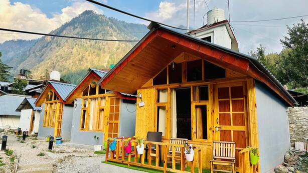 A row of wooden cabins with a mountain backdrop, featuring a porch with chairs and potted plants. - The Aisling House (Bhuntar)
