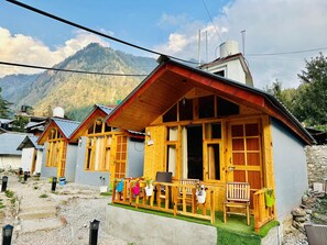 A row of wooden cabins with a mountain backdrop, featuring a porch with chairs and potted plants. - The Aisling House (Bhuntar)