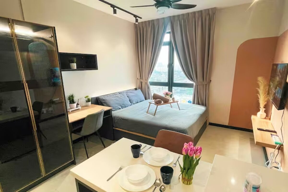 Une chambre d’hôtel moderne, équipée d’un lit, d’un bureau et d’une chaise. On y trouve une fenêtre avec des rideaux, un ventilateur de plafond et une petite table sur laquelle est posé un vase de fleurs.