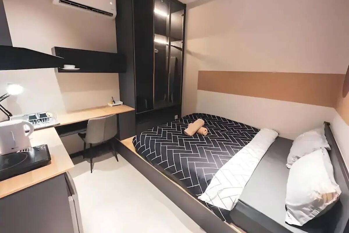 A compact hotel room with a bed, a desk, a chair, a wardrobe, and a ceiling fan.