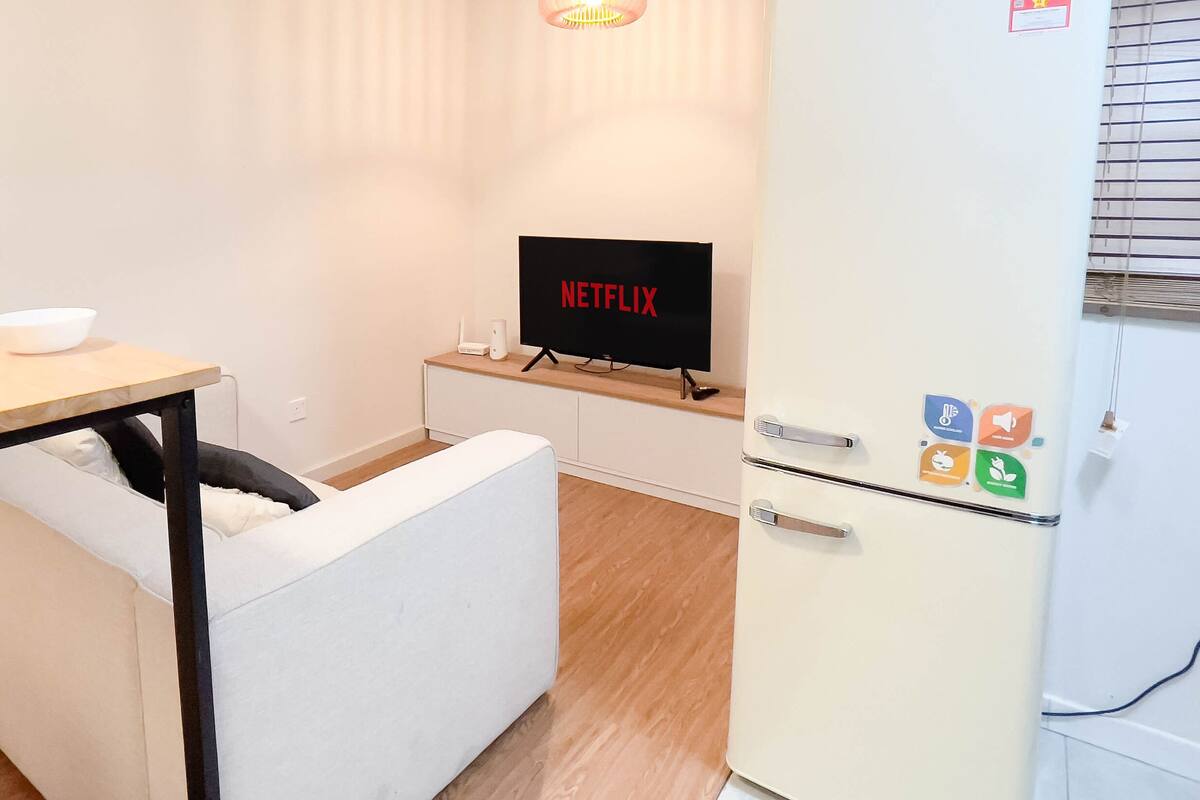 A compact kitchenette with a microwave, a small refrigerator with a washing machine, a dining table with white plates and glasses, and a TV displaying the Netflix logo.