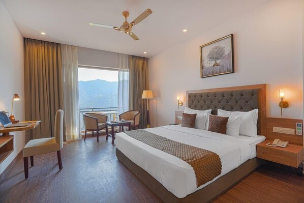 Hotelkamer met een groot bed, een bureau met stoel, een plafondventilator en uitzicht op de bergen door het raam.