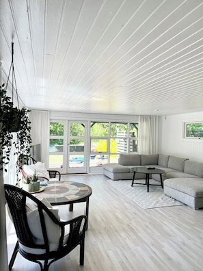 A modern living room with a wooden ceiling, a grey sectional sofa, a round coffee table, and a dining area with a table and chairs. - Charming summer house on a secluded plot (Glesborg)