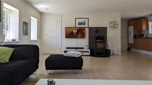 A modern living room with a black sofa, a white ottoman, a flat-screen TV, a fireplace, and a kitchen area. - Cozy country estate in Vesterborg. (Vesterborg)