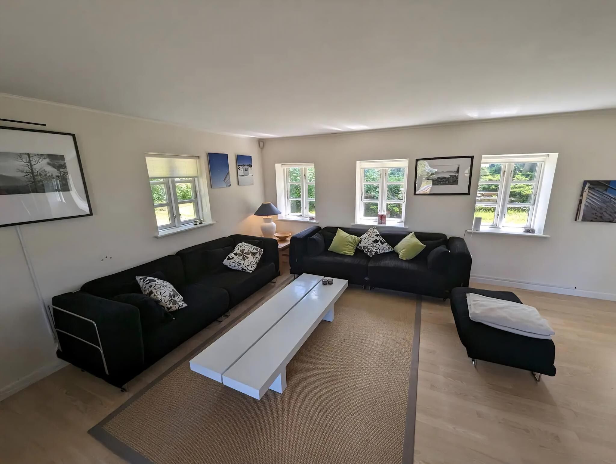 A modern living room with a sectional sofa, a white bench, and a small table. There are multiple windows providing natural light.