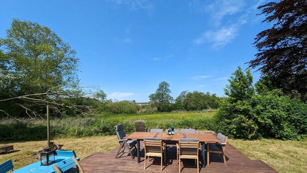 An outdoor wooden deck with a dining table and chairs, surrounded by greenery and trees. - Cozy country estate in Vesterborg. (Vesterborg)