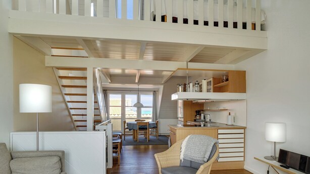 Un intérieur moderne comprenant un canapé, une table à manger, une chaise en osier, un coin cuisine et un escalier menant à un niveau supérieur.