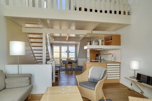 A modern interior with a sofa, dining table, wicker chair, kitchen area, and a staircase leading to an upper level.