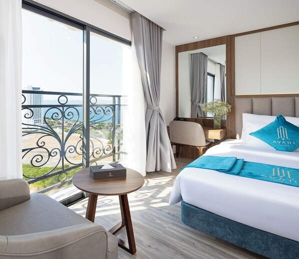 A hotel room with a bed, a desk, a chair, a small table, and a balcony with a view of the sea. - Avari Hotel (Nha Trang)