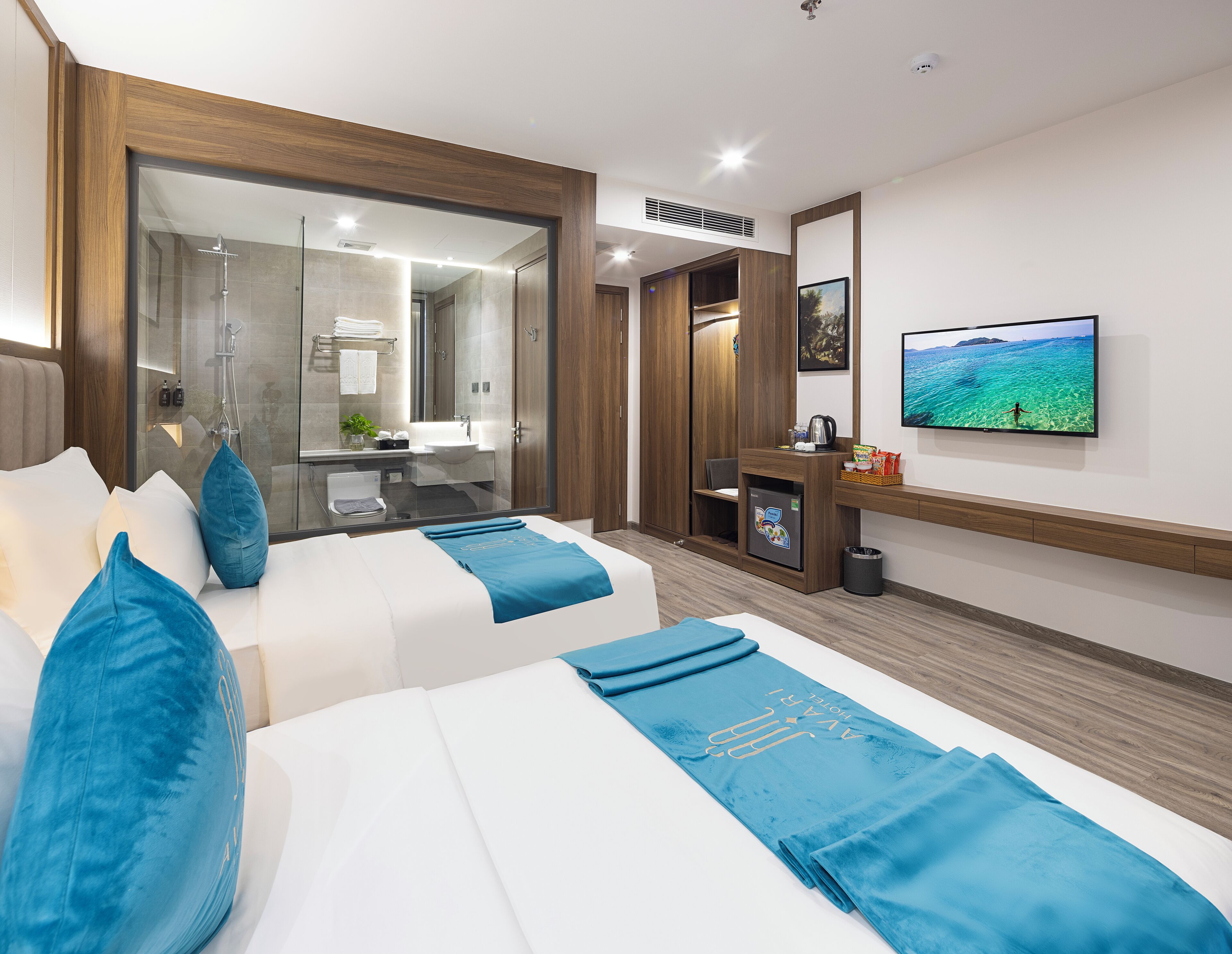 A hotel room with two beds, a TV, a desk, a mirror, and a bathroom visible through a glass door.