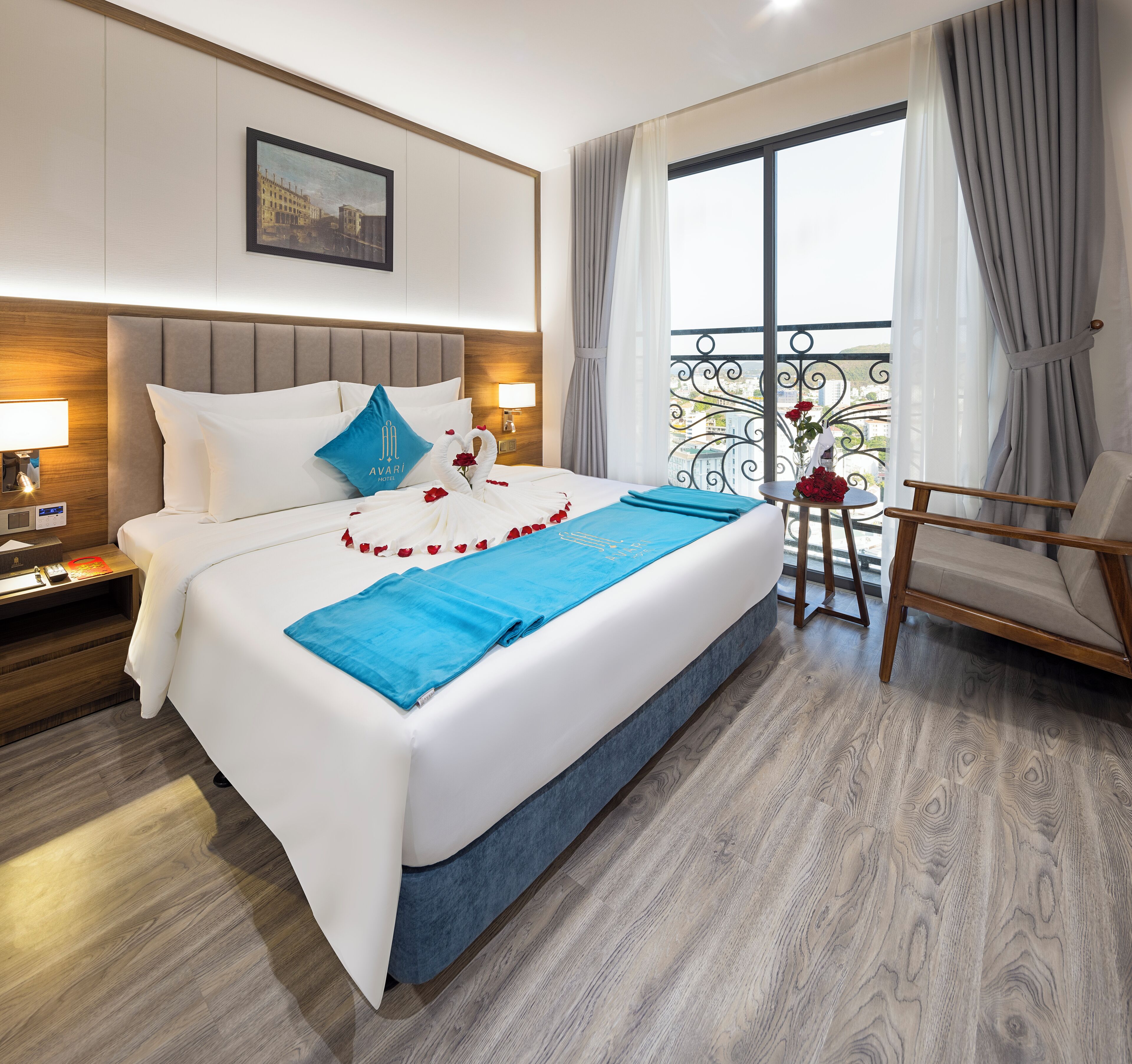 Habitación de hotel con una cama grande, una mesita de noche con lámpara, vistas de la ciudad desde un balcón y un cuadro en la pared.