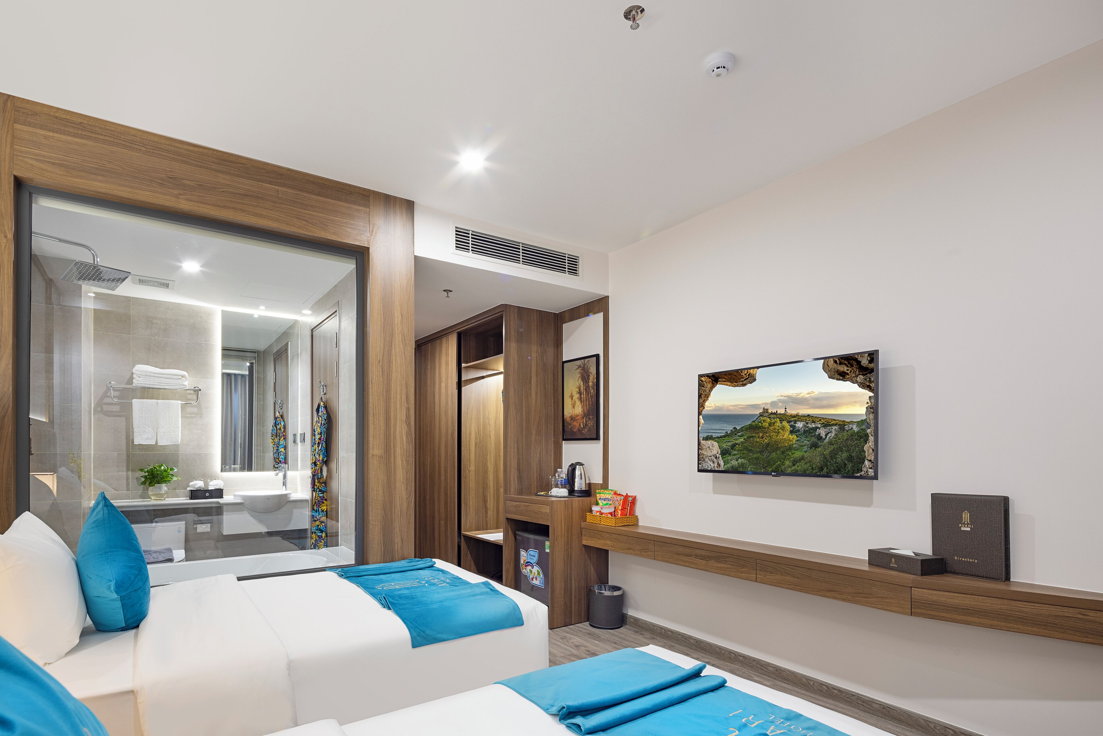 Une chambre d’hôtel moderne avec une grande fenêtre, un lit avec une literie bleue, un petit bureau et un tableau représentant un paysage côtier accroché au mur.