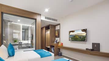 Une chambre d’hôtel moderne avec une grande fenêtre, un lit avec une literie bleue, un petit bureau et un tableau représentant un paysage côtier accroché au mur.