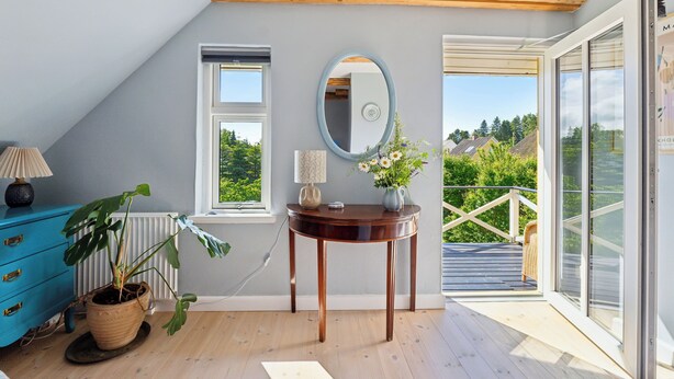A room with a wooden ceiling, a large window, a mirror, a table with a vase, and a blue dresser.