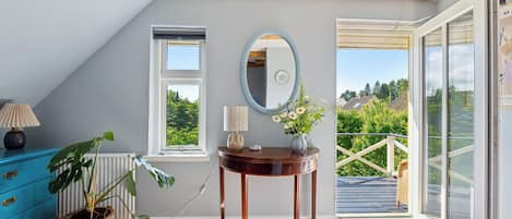 A room with a wooden ceiling, a large window, a mirror, a table with a vase, and a blue dresser.
