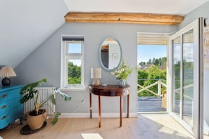 Una habitación con techo de madera, un ventanal grande, un espejo, una mesa con un jarrón y un aparador azul.