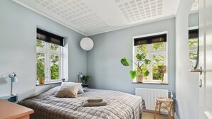 A bedroom with a bed, two windows with blinds, a round ceiling light, a radiator, and a small wooden stool.