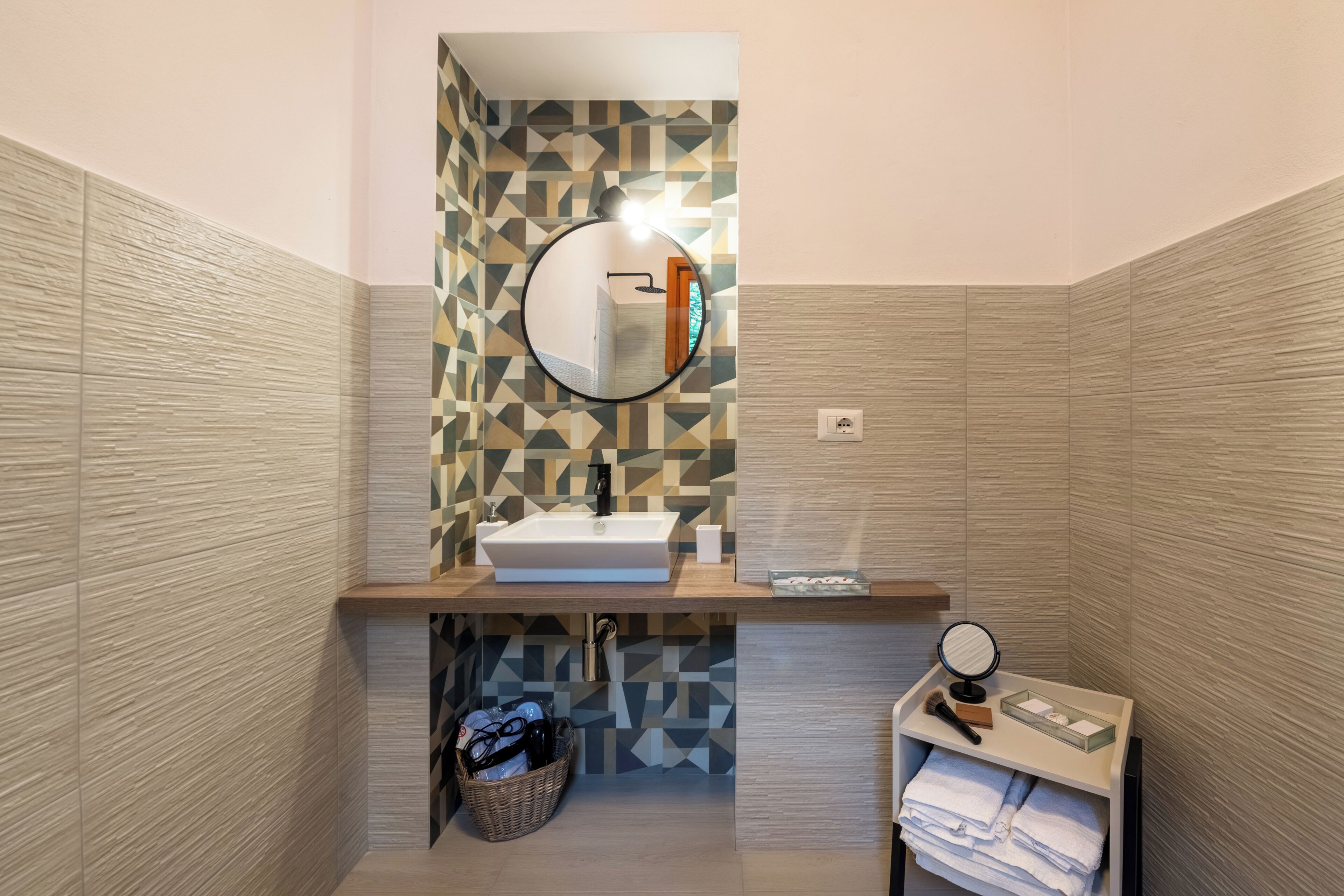 Un baño moderno con un muro de diseño geométrico, un lavamanos con espejo y una pequeña tocador con toallas y artículos de aseo.