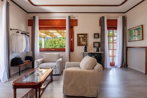 Uma sala de estar bem iluminada, com sofá, poltrona e mesa de centro. Há uma janela com cortinas, um quadro na parede e um pequeno armário.