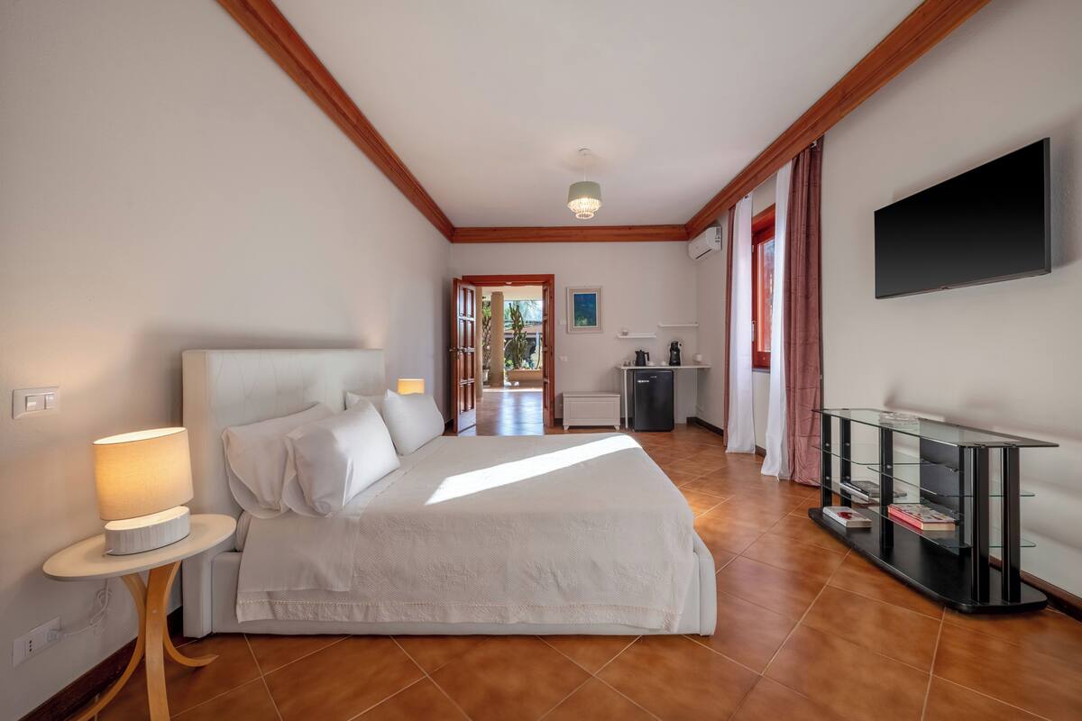 Quarto com cama, mesa de cabeceira, abajur e televisão na parede.