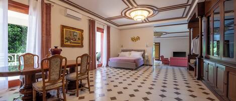 Une chambre d’hôtel avec un grand lit, une table à manger avec des chaises, un lustre et une vue sur la verdure.