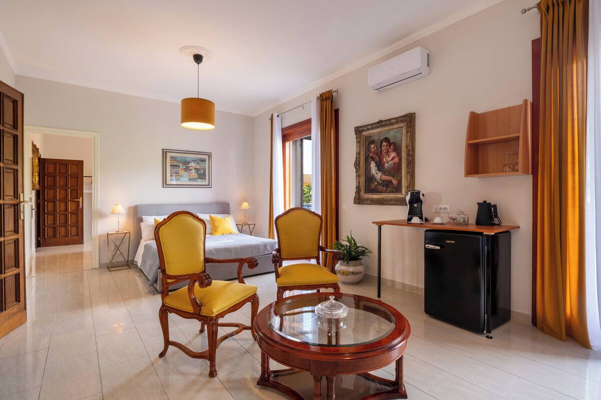 Quarto de hotel com uma cama, duas cadeiras, uma mesinha, um refrigerador, uma planta em vaso e um quadro na parede.