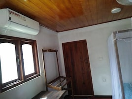 Quarto com teto de madeira, janela com moldura de madeira, ar condicionado branco e porta de madeira, além de uma cama com colcha branca.