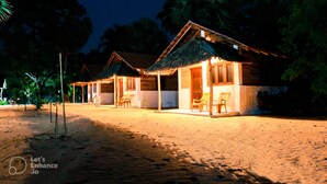 A row of thatched-roof cottages with outdoor seating, set on a sandy beach at night. - Ocean View Beach Resort (Kudawa)