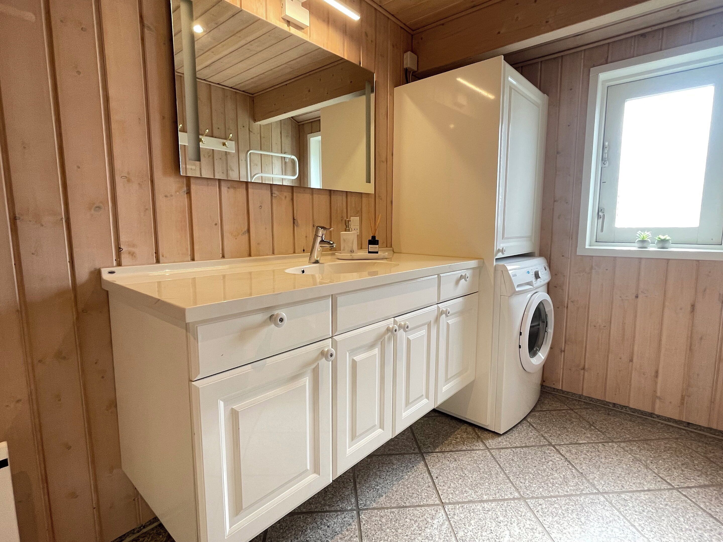Eine Küche mit Holzwänden, einer weißen Arbeitsplatte, einem Spülbecken, einem Spiegel, einer Waschmaschine und einem Fenster mit Pflanzen.