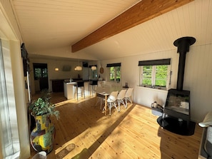 A spacious room with a wooden floor, a dining area with chairs, a kitchen with a stove, and a large window. - Big renovated cottage with soul and love (Grevinge)