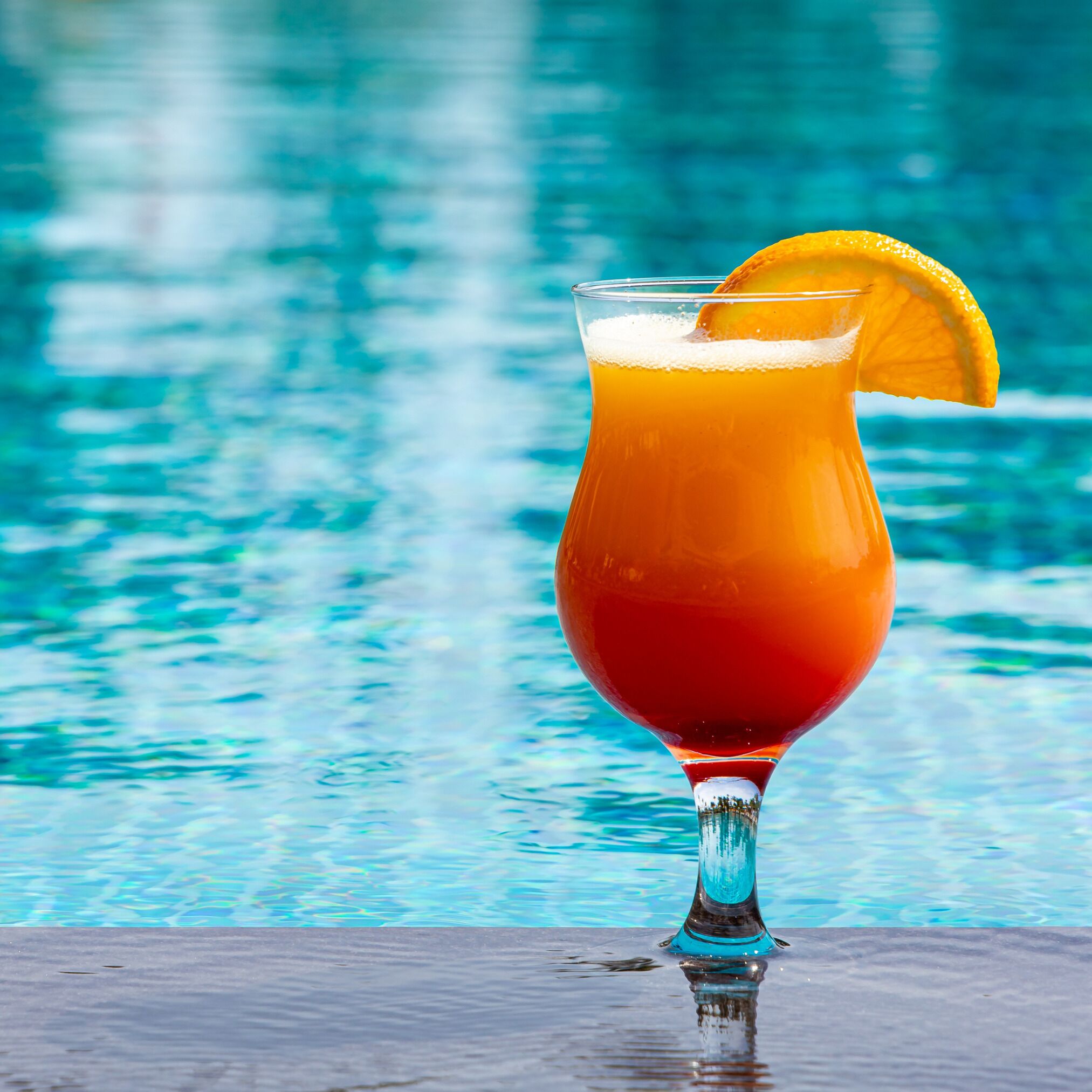 Un cocktail servi dans un verre, agrémenté d’une tranche d’orange, posé sur une surface près d’une piscine.