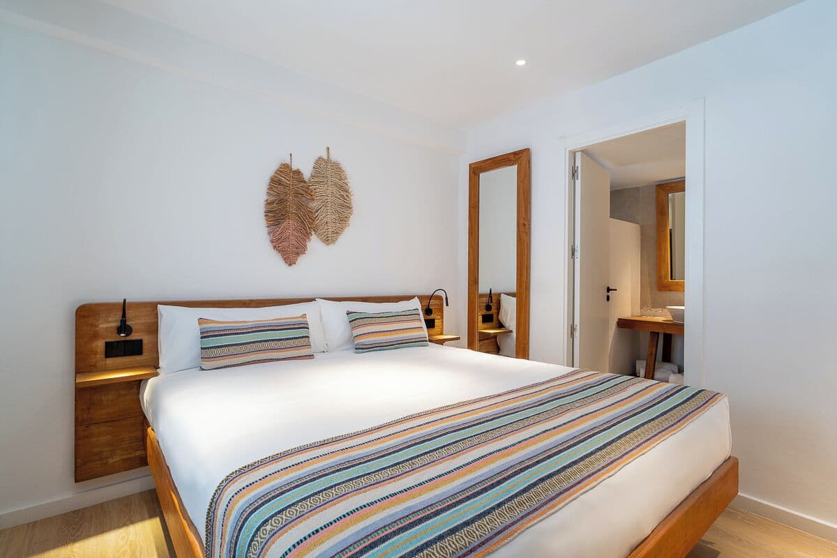 A bedroom with a large bed, a wooden headboard, a bedside table with a lamp, and a wall-mounted mirror.