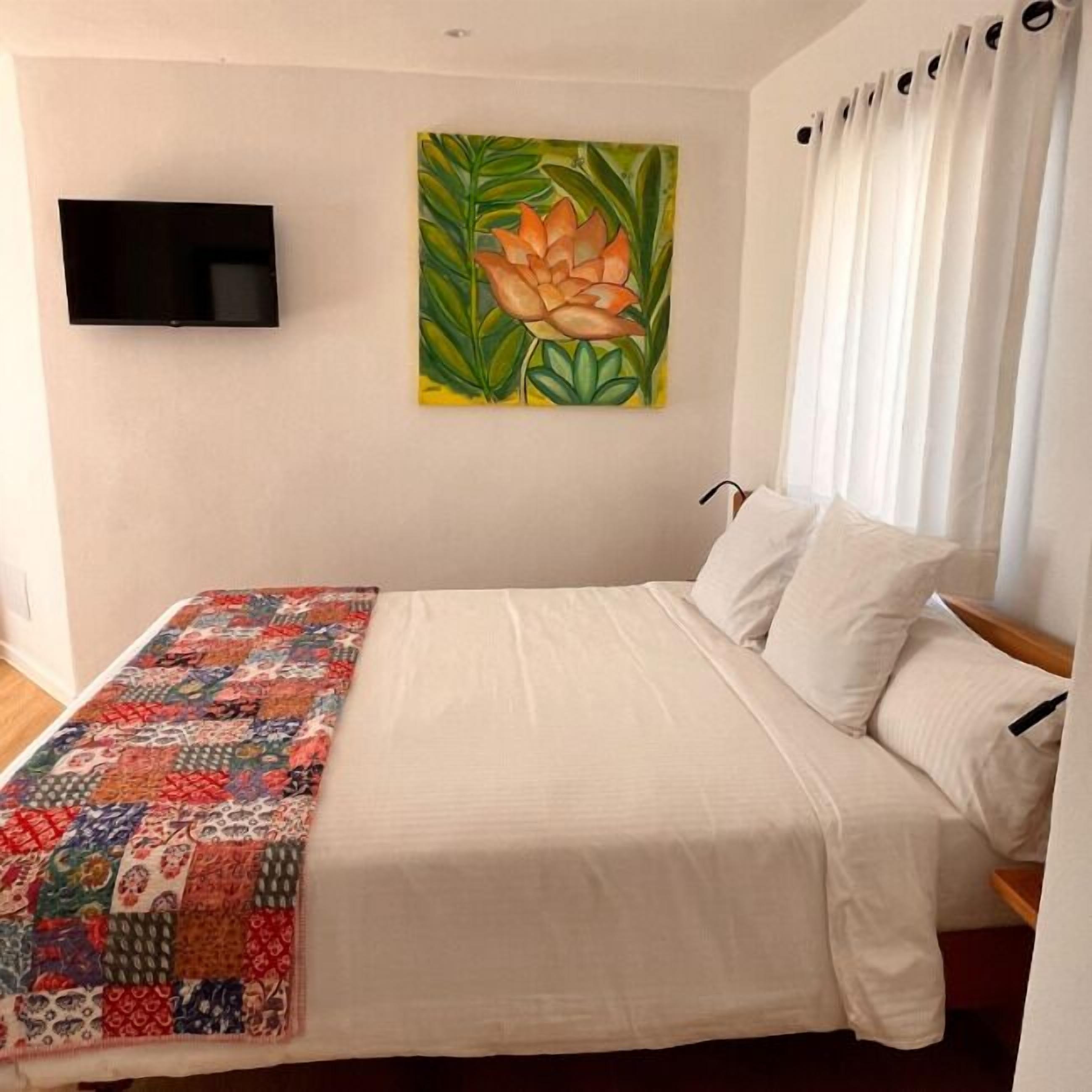 A bedroom with a bed, a quilt, a painting, and a TV on the wall.