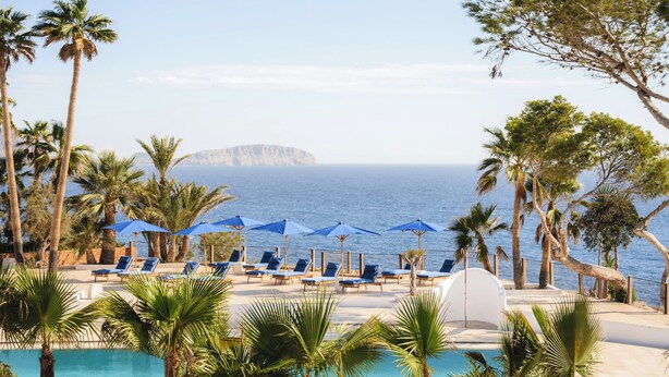 A poolside area with lounge chairs and umbrellas, palm trees, and a view of the sea and a distant island.