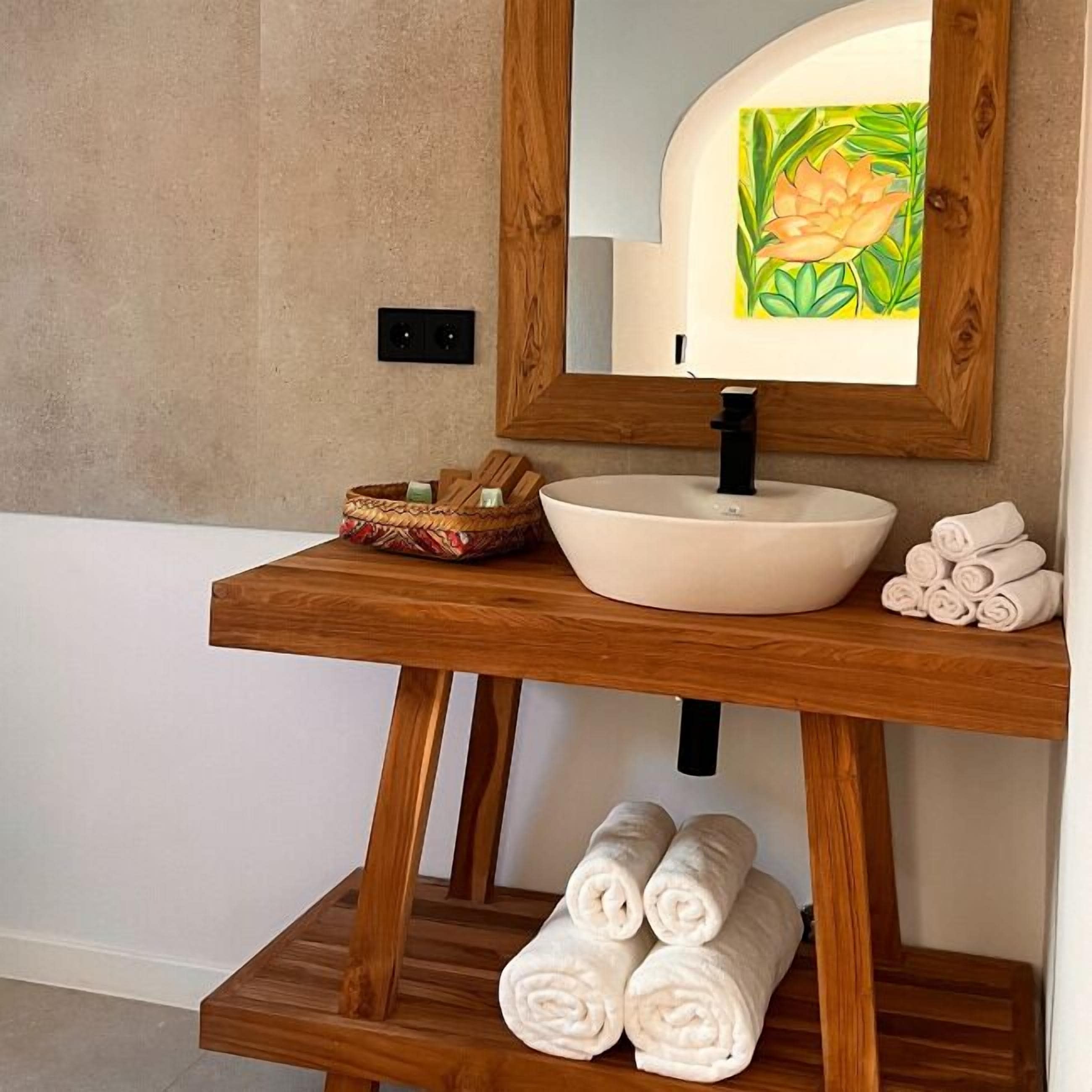 A bathroom with a wooden vanity, a white sink, a mirror with a floral frame, and a shelf with rolled towels.
