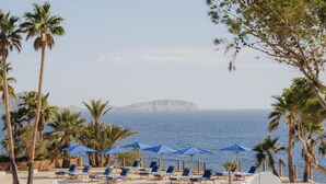 A beachside lounge area with sun loungers and umbrellas, palm trees, and a view of the sea and a distant island.