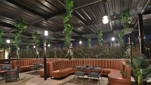 A modern lounge area with a curved brown leather sofa, black wicker tables, and hanging green plants.