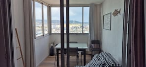 Una habitación pequeña con vista a un puerto deportivo y colinas. Cuenta con una mesa, dos sillas, un sofá y una escalera.