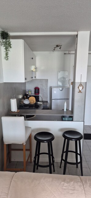 Cocina pequeña con mostrador, cafetera y lavamanos. Hay dos taburetes y una silla en el comedor.