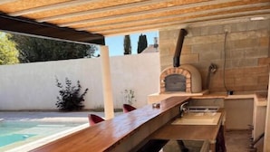 A wooden bar counter with a built-in sink, a stone fireplace, and a pool in the background. - 1 Bedroom Apartment in Beauvoisin (Beauvoisin)