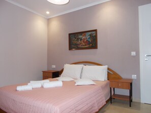 A neatly made bed with pink bedding and white pillows, a wooden nightstand with a lamp, and a painting on the wall. - ΜΙΚΡΟ ΣΠΙΤΑΚΙ (Κοζάνη)