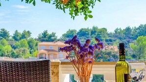 A table with a bottle of olive oil and a glass, a vase with purple flowers, and a wicker chair. - Apartments Katica's Place (Blato)