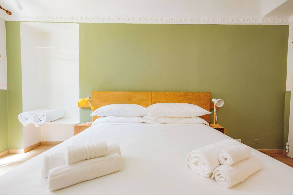 A hotel room with a bed, white linens, a wooden headboard, a nightstand, and a lamp.